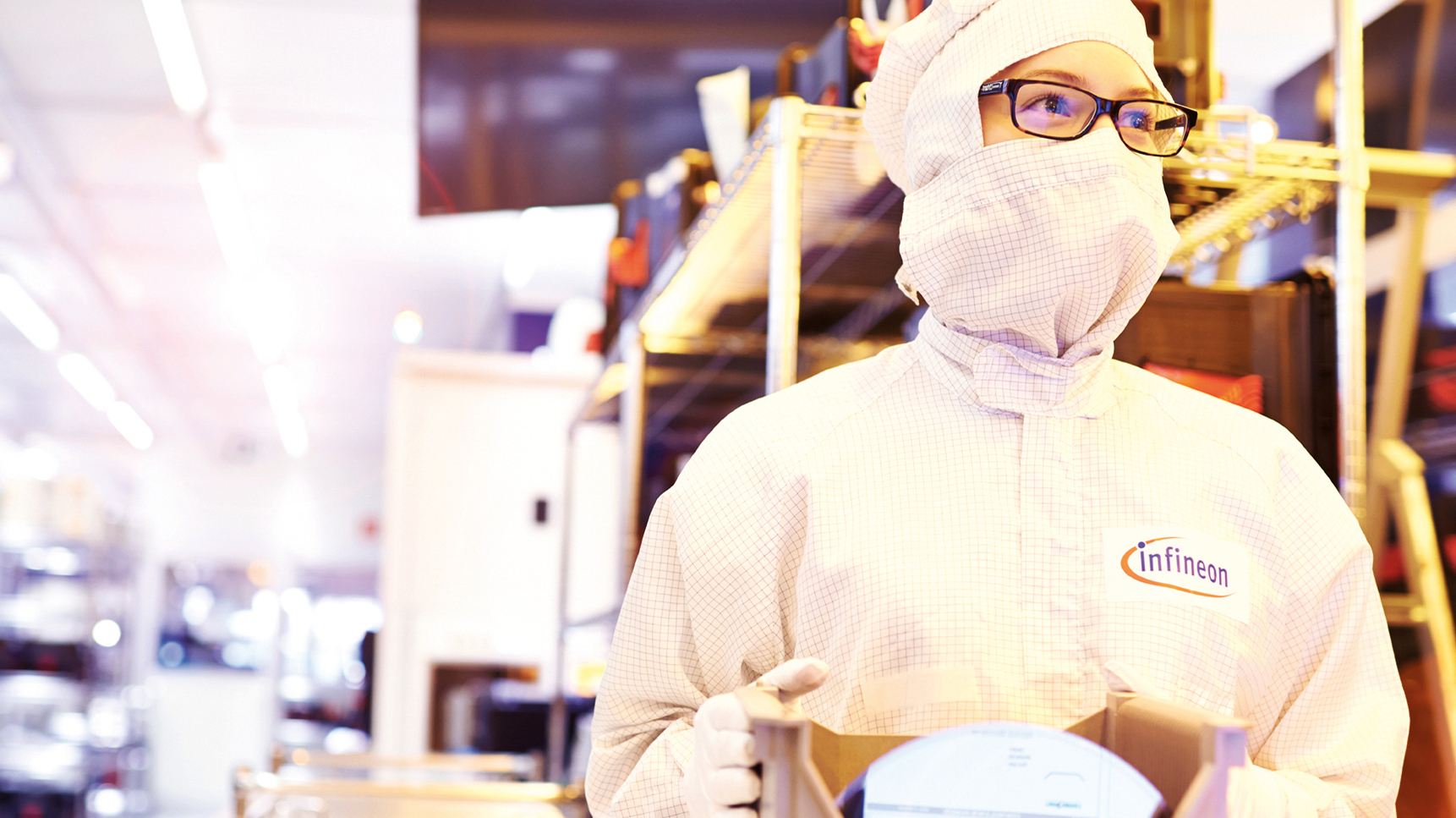 Eine Person in Reinraumkleidung steht in einem hell beleuchteten Labor und hält ein technisches Gerät in den Händen. Auf dem Anzug steht „Infineon“.