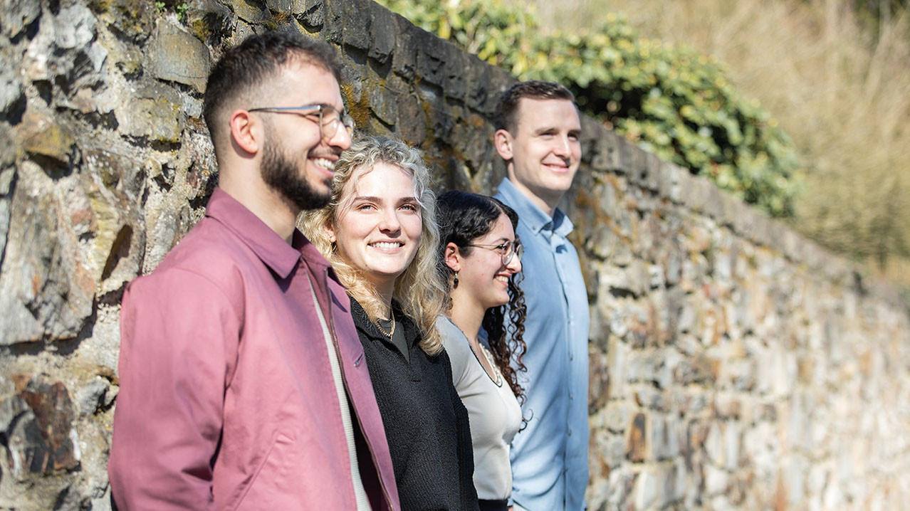 4 Personen stehen schräg vor einer alten Steinmauer.