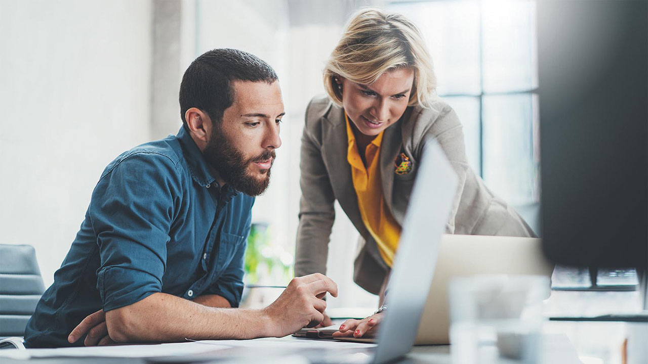 Zwei Personen arbeiten gemeinsam an einem Laptop: Eine Person sitzt und tippt, die andere beugt sich danebenstehend über den Bildschirm.