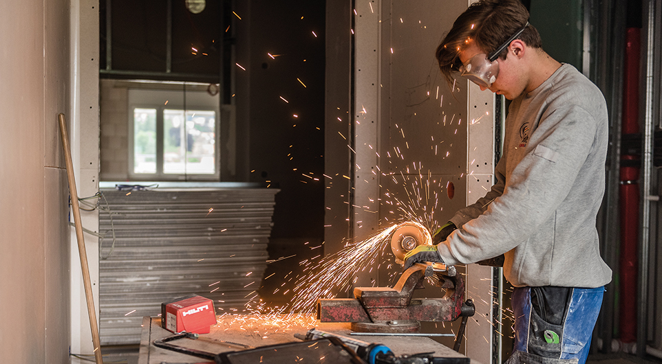 Junger Mann beim Schleifen an einem Werkstück. Der junge Mann arbeitet in einem Rohbau, er trägt eine Schutzbrille und die Funken fliegen am Werkstück.