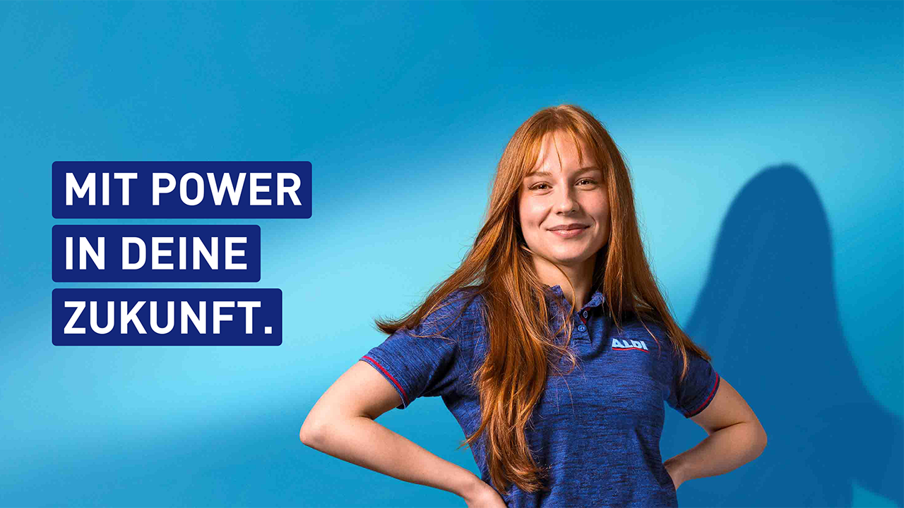 Eine Person in blauem T-Shirt steht vor einem blauen Hintergrund, lächelt und wirkt selbstbewusst. Daneben steht der Slogan: MIT POWER IN DEINE ZUKUNFT
