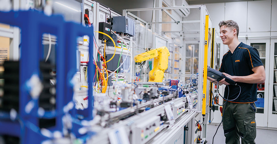 Eine Person steht in einem Technikraum und bedient eine Steuerungseinheit. Vor ihr befindet sich eine lange Werkbank mit vielen Kabeln, Geräten und einem gelben Roboterarm.