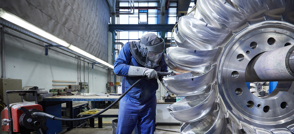Das Bild zeigt eine Person in Schutzkleidung bei der Bearbeitung eines großen metallischen Turbinenrades in einer Werkhalle.