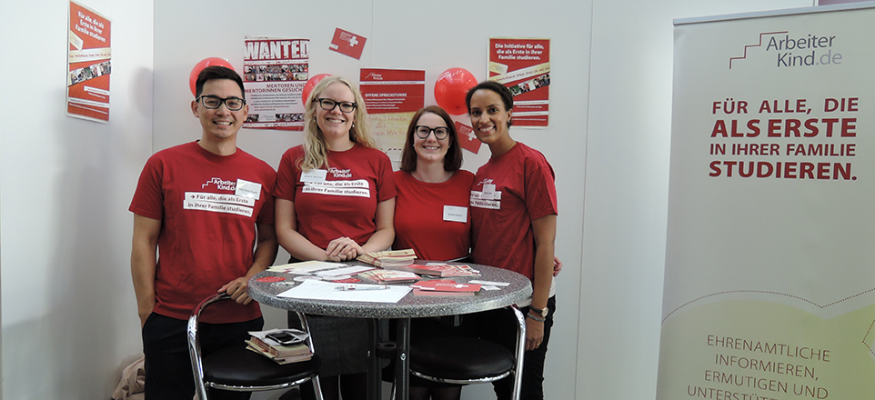 Vier Personen in roten T-Shirts stehen an einem Messetisch. Daneben steht ein Roll-Up von ArbeiterKind.de mit der Aufschrift: FÜR ALLE, DIE ALS ERSTE IN IHRER FAMILIE STUDIEREN.