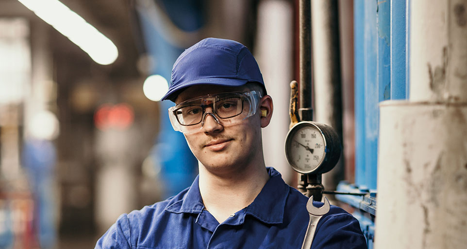 Eine Person in blauer Arbeitskleidung, Schutzbrille und Mütze steht mit verschränkten Armen in einer Fabrikhalle. Neben ihr ist eine große Maschine mit einem Druckmesser.