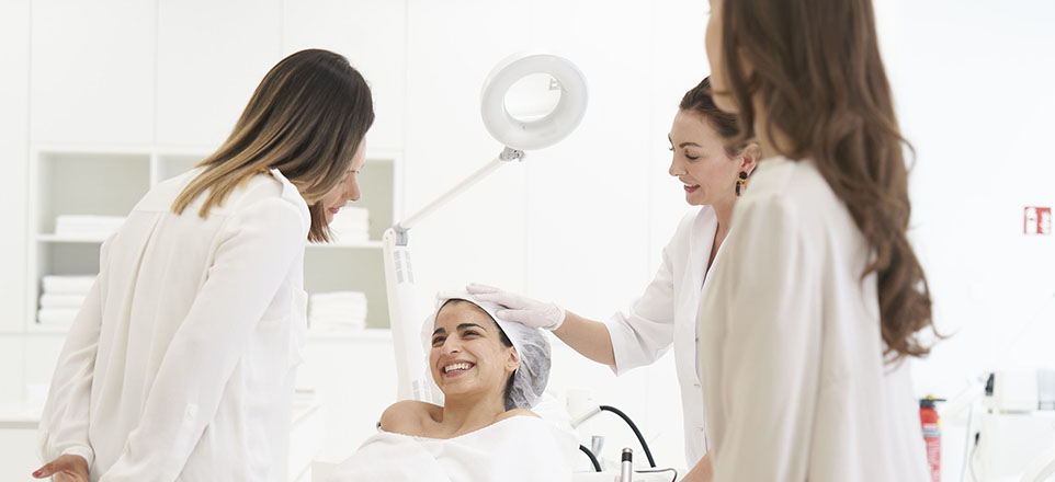 3 Frauen mit weißen Blusen kümmern sich um eine Patientin die mit einer weißen Haarhaube auf einer Behandlungsliege liegt