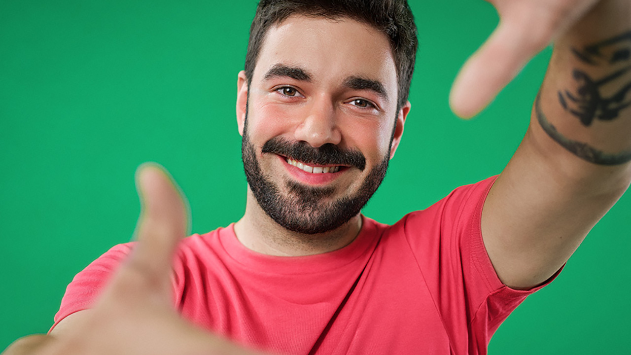 Eine Person steht vor einem grünen Hintergrund, lächelt und hält die Hände wie einen Rahmen vor sich.