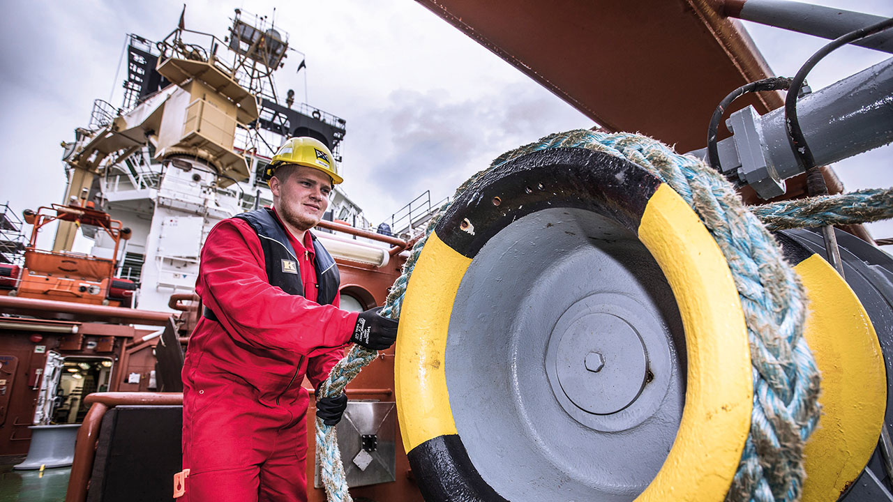 Person in roter Arbeitskleidung mit Helm zieht an einem dicken Tau, Containerschiff im Hintergrund