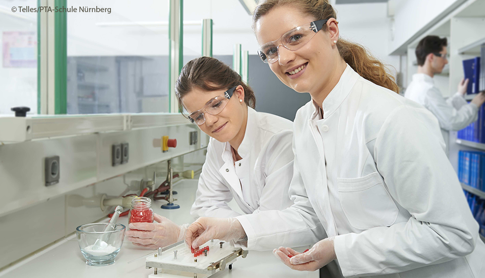 Zwei Pharmazeutisch-technische Assistent:innen (PTA) bei Ihrer Arbeit im Labor in Kitteln und mit Schutzbrillen