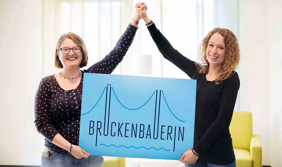 Zwei Frauen bauen mit den Armen eine Brücke und halten ein Schild "Brückenbauerin"
