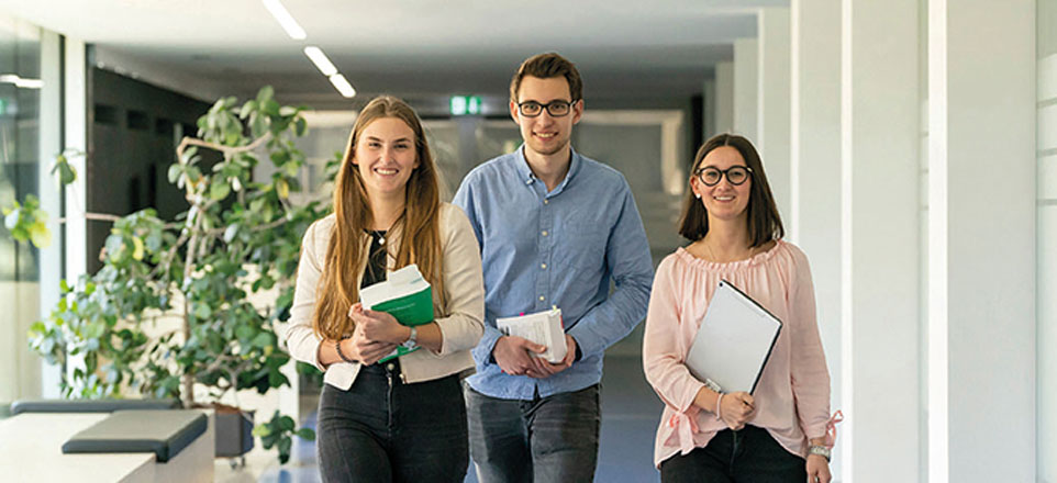 Drei Personen gehen einen Gang entlag und halten Akten oder Bücher in den Händen.