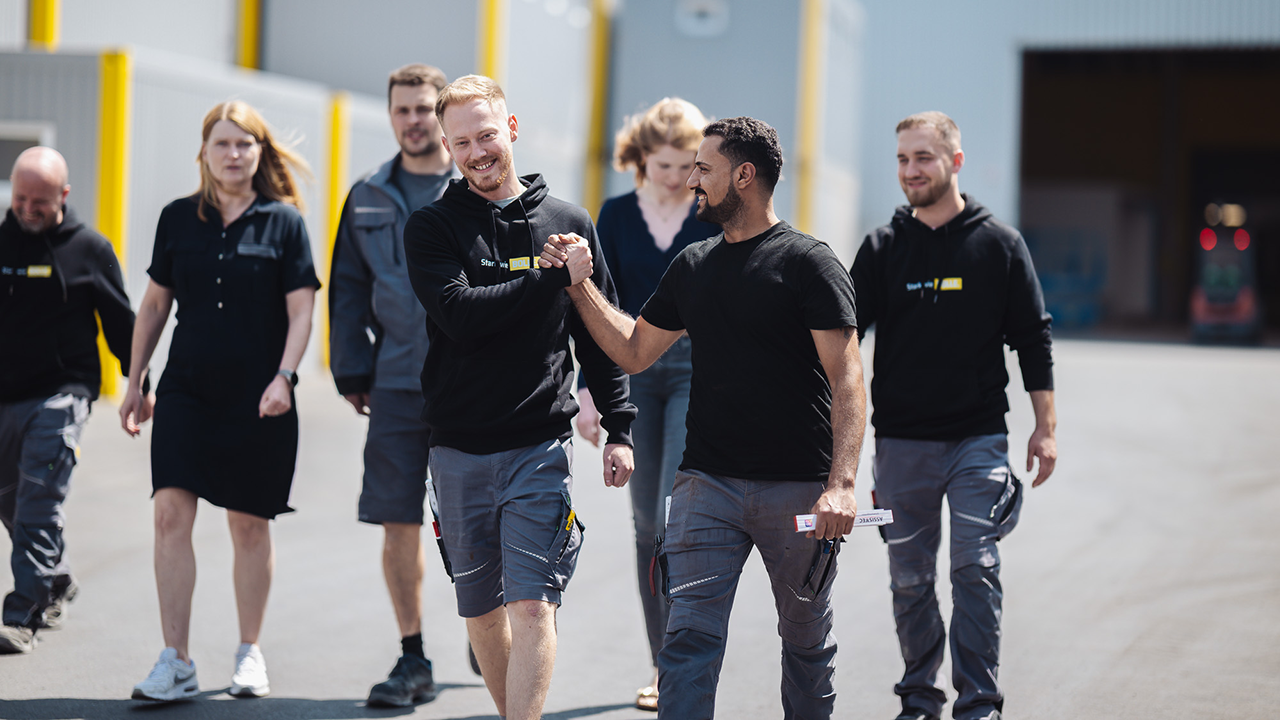 Foto von sieben Mitarbeitenden die auf dem Firmengelände nebeneinander herlaufen. Die zwei vorderen klatschen sich gerade ab.