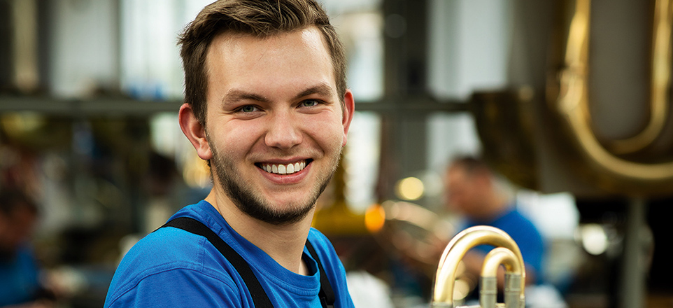 Eine junge Person in Arbeitskleidung sitzt lächelnd in einer Werkstatt, umgeben von Blasinstrumenten.
