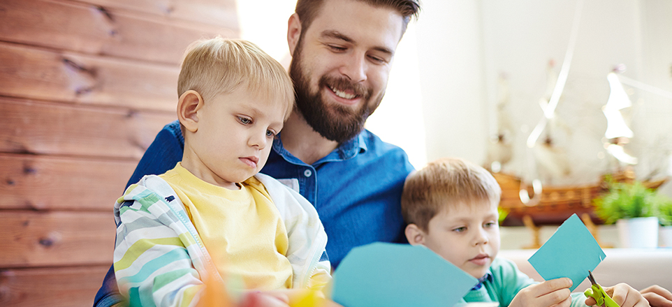 Zwei Kinder sitzen auf dem Schoß einer erwachsenen Person und basteln mit Schere und Papier.