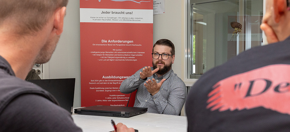 Es sind drei Personen zu sehen, eine von ihnen frotal. Diese erklärt mithilfe von Gesten des anderen beiden etwas. 
