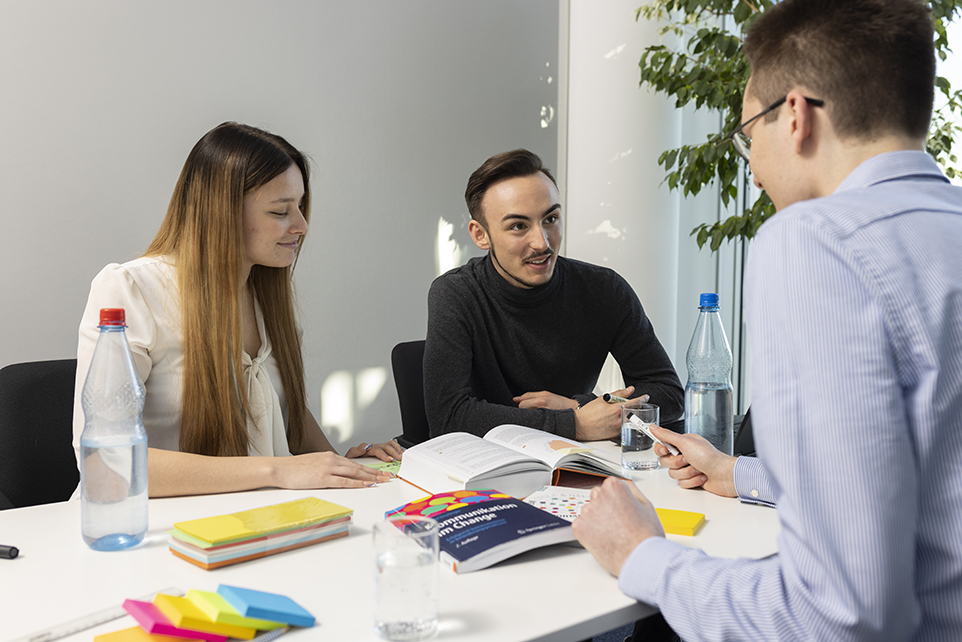 Drei Personen sitzen an einem Tisch und unterhalten sich. Auf dem Tisch liegen Bücher und weitere Arbeitsmaterialien..