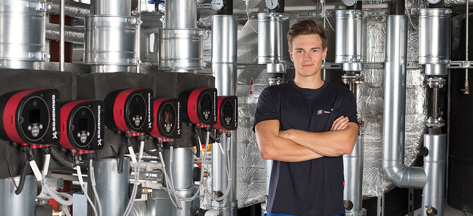 Junge Person mit verschränkten Armen vor einer Anlage für Gebäudetechnik