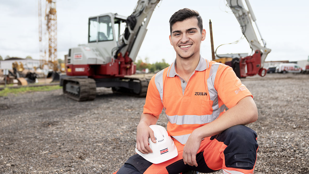 Junger lächelnder Mensch in orangefarbener Schutzkleidung mit Helm in der Hand kniet vor einem Bagger auf einer Baustelle