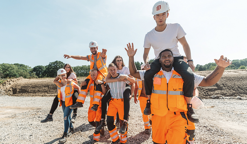 Bild von mehreren jungen Leuten in Arbeitskleidung die sich jeweils Huckepack nehmen. 