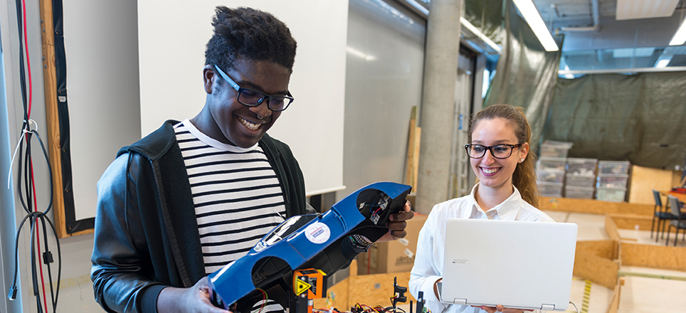Eine lächelnde Person mit Brille mit einem Modellauto in der Hand und eine lächelnde Person mit Brille mit einem Laptop in der Hand