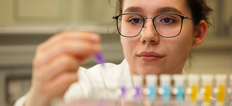 Eine Person im Laborkittel hält konzentriert eine Pipette über bunte Flüssigkeiten in kleinen Reagenzgläsern.
