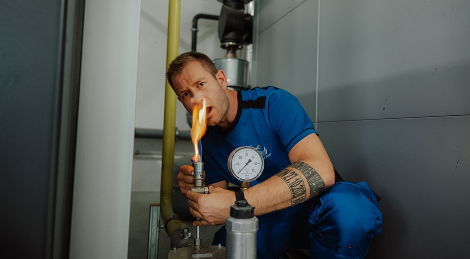 Handwerker in blauer Arbeitskleidung. Er hält ein Prüfgerät oder Ventil in der Hand, aus dem eine offene Flamme austritt – vermutlich wird hier eine Brenner- oder Dichtheitsprüfung durchgeführt. Direkt daneben ist ein Druckmanometer zu sehen.