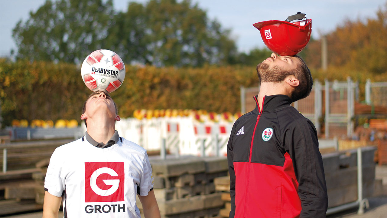 Zwei Personen, eine balanciert einen Fußball auf dem Kopf, ein anderer einen Bauhelm.