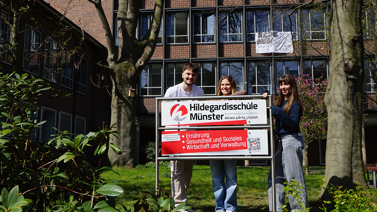 Drei Schüler/innen stehen hinter dem Schulschild vor dem Schulgebäude.