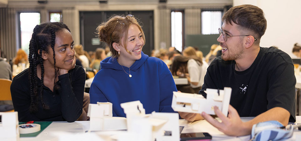 Drei Personen sitzen an einem Tisch, unterhalten sich, lächelnd und halten Architekturmodelle in den Händen. 