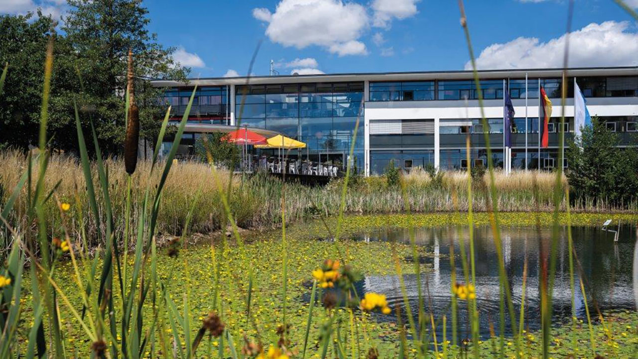 Aufnahme der Hochschule Hof. Im Vordergrund ist ein Teich zu sehen.