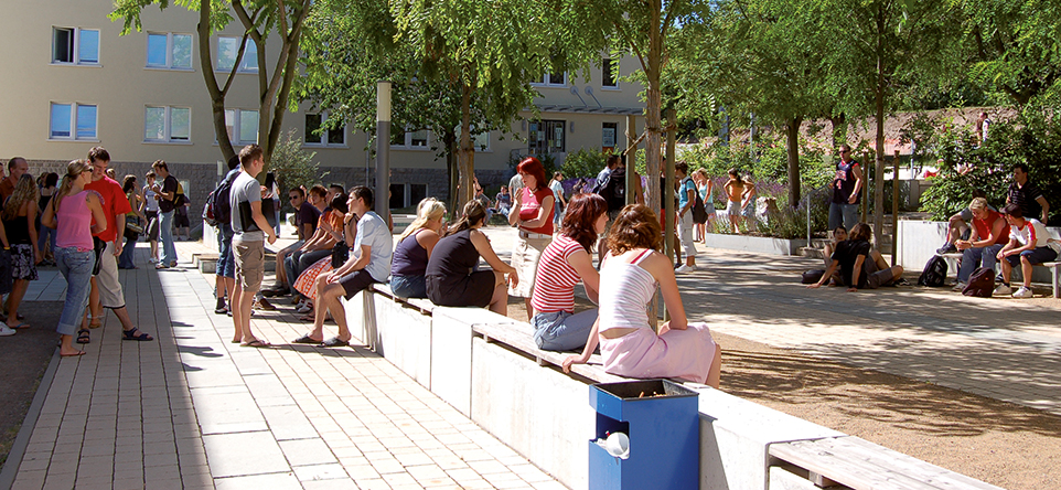 an einem sonnigen Tag sitzen und stehen Studierende im Aussenbereich der Hochschule in verschiedenen Gruppen zusammen