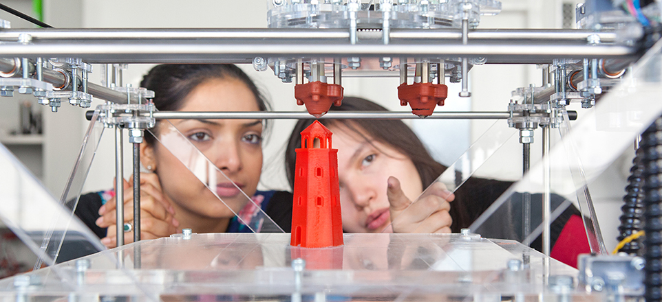 Großaufnahme eines 3D Druckers, beim Drucken eines roten Turmes im Hintergrund zwei Mitarbeiterinnen, die sich darüber austauschen