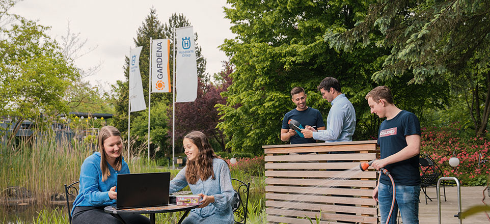 Fünf junge Personen auf einem Gartengelände mit kleinem Teich, zwei sitzen am Laptop, einer bewässert den Garten, zwei sind im Gespräch