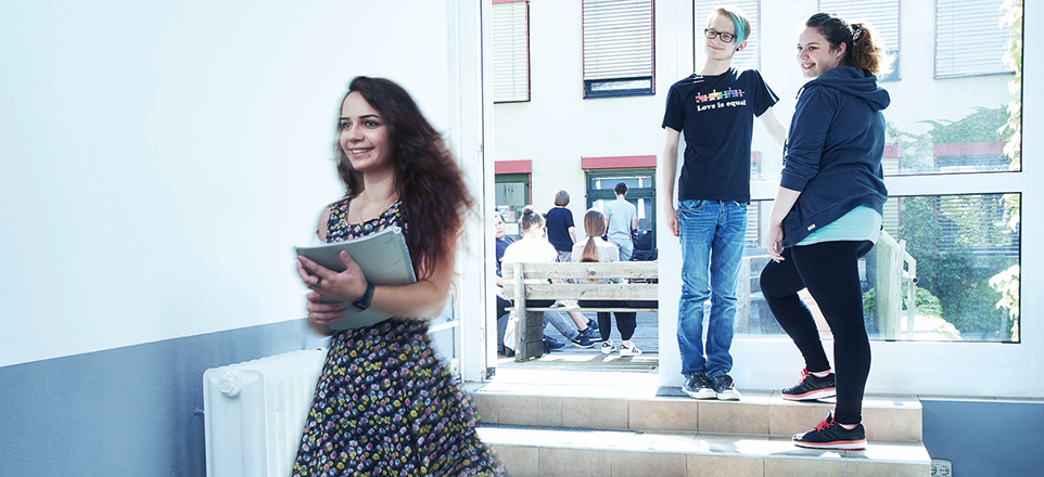 Eine Person läuft mit einem Block durch den Flur eines Gebäudes, zwei andere stehen auf einer Treppe, weitere sitzen draußen auf Bänken.