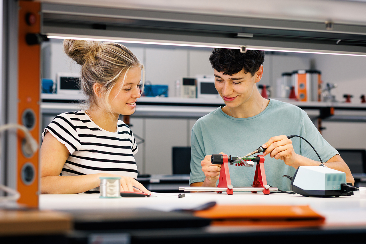 Zwei Personen sitzen in einer Werkstatt an einem Tisch. Eine Person hält einen Lötkolben und arbeitet an einem kleinen Bauteil, die andere schaut interessiert dabei zu.