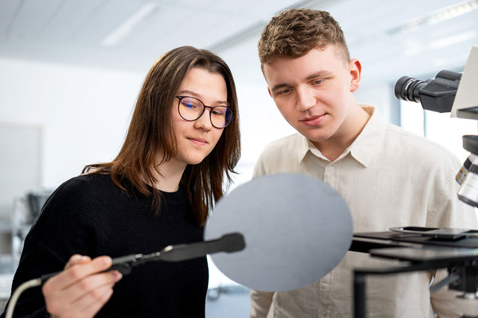 Zwei junge Personen stehen neben einem Mikroskop und begutachten eine Scheibe