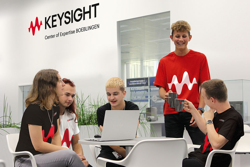 Fünf junge Menschen sitzen/stehen um einen Laptop, der auf einem Tisch steht und unterhalten sich.
