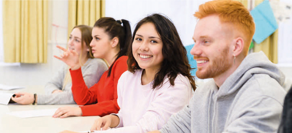 4 junge Menschen im Klassenzimmer an Tischen sitzend.
