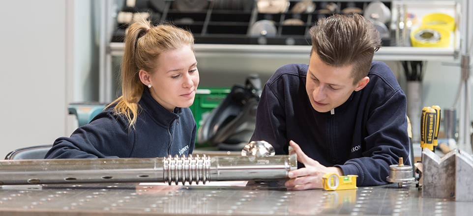 Zwei Personen prüfen gemeinsam ein Metallbauteil auf einem Werkstatttisch, umgeben von Werkzeugen.