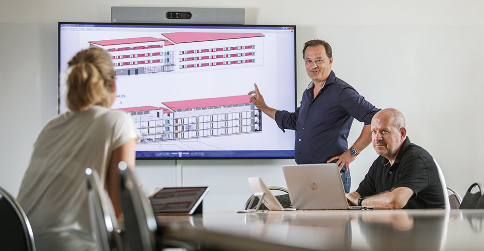 Drei Personen besprechen Architekturpläne, die auf einem großen Bildschirm angezeigt werden.