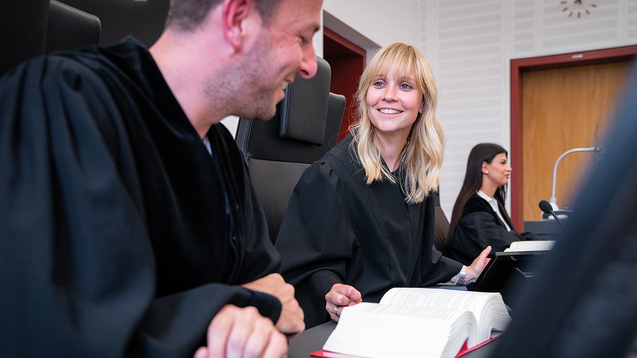 Zwei lächelnde Personen im Gericht mit einem Gesetzesbuch, im Hintergrund eine weitere Person