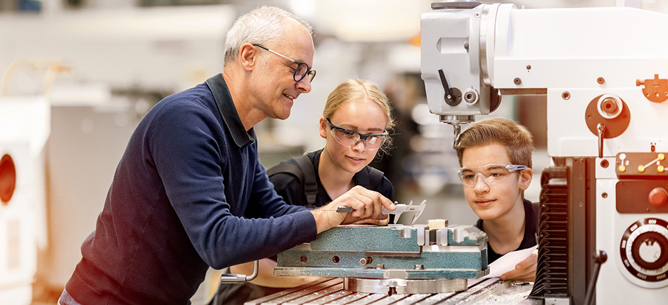 Drei Personen stehen an einer großen Maschine in einer Werkstatt. Eine Person erklärt etwas, die anderen hören aufmerksam zu. Alle tragen Schutzbrillen.