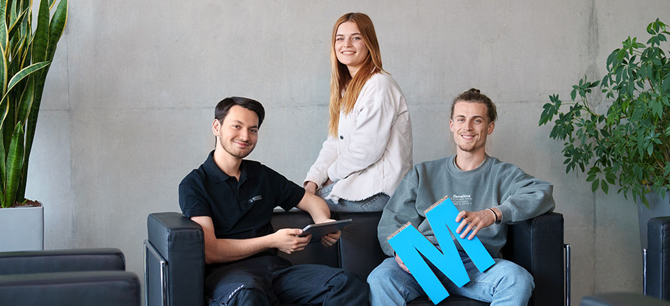 Drei Personen sitzen auf einer Couch, eine hält ein blaues M.