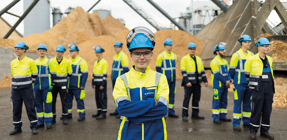 Gruppenbild: mehrere Personen in gelb-blauen Arbeitsnzügen mit hellblauem Schultzhelm stehen vor einem großen Sandberg