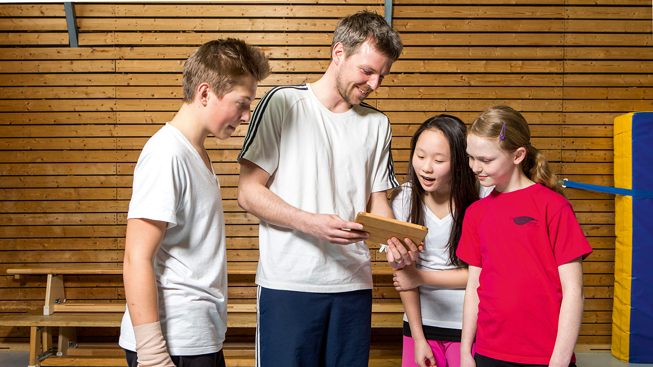 Lehrer zeigt Schülerinnen und Schülern etwas auf dem Tablet in der Sporthalle