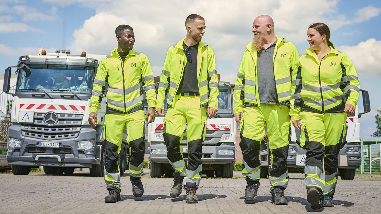 Vier Mitarbeitende in gelber Arbeitskleidung, im Hintergrund drei LKWs