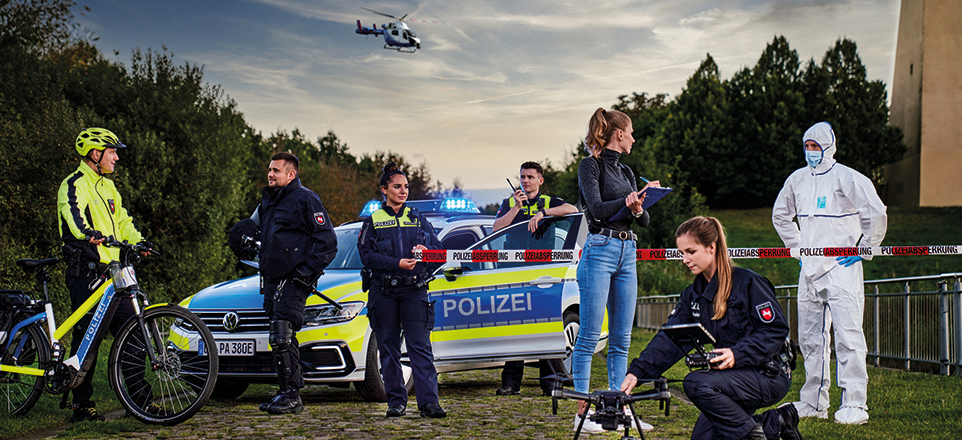 Gruppenfoto von sieben Menschen aus verschiedenen Bereichen der Polizeidirektion Osnabrück, im Hintergund ein Helikopter am Himmel
