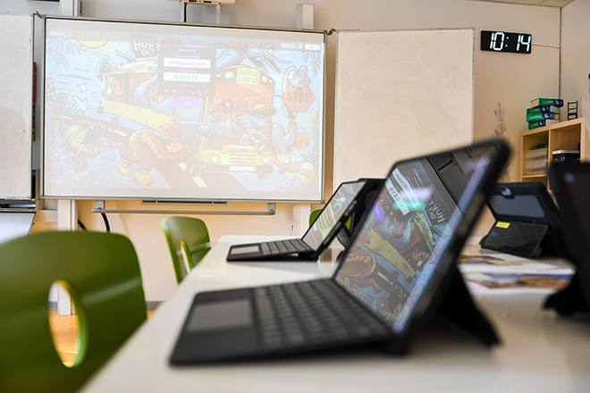 Szene aus einem Klassenraum: Ein Tisch auf dem aufgeklappte Tablets stehen. Im Hintergrund sieht man eine digitale Tafel.