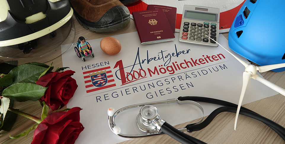 Verschiedene Gegenstände wie Helm, Rose, Stethoskop, Reisepass, Spielzeug, Taschenrechner und Baustiefel liegen um ein Plakat des Regierungspräsidiums Gießen mit dem Slogan „1 Arbeitgeber, 1000 Möglichkeiten“.