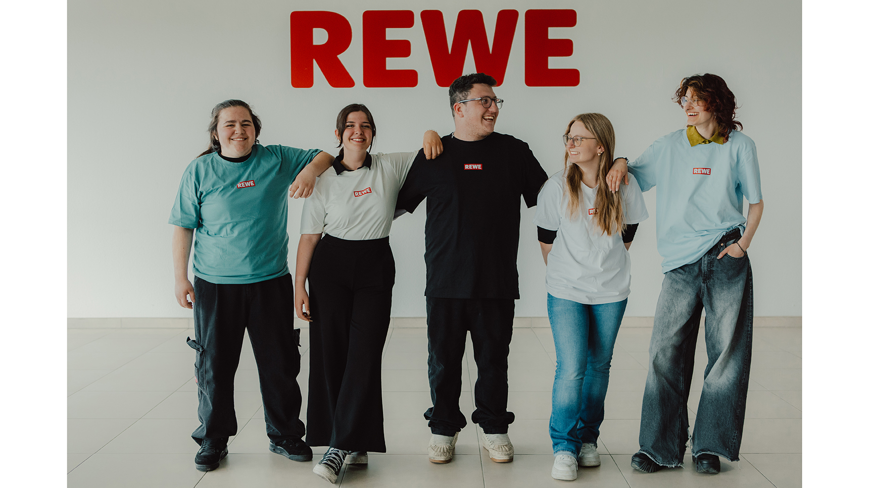 Gruppe von 5 Menschen die sich in den Armen locker halten. Sie stehen vor einem REWE Schild